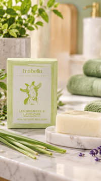 Frabella Lemongrass & Lavender goat milk soap bar with its light green packaging box, styled on a marble countertop with green herbs, a bar of soap on a marble dish, and green towels in the background.