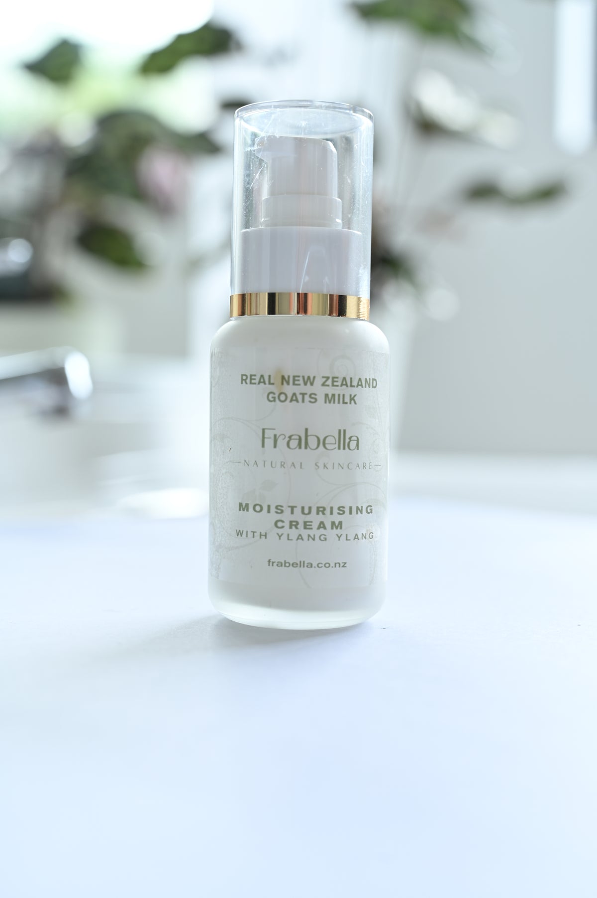 A pump bottle of Ylang Ylang Moisturising Cream with Kawakawa on a white surface, with a plant in the background.
